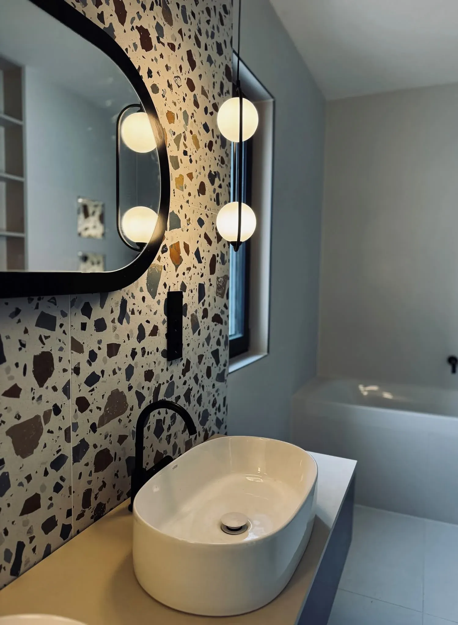 Fürdőszobai mosdó ovális tükörrel, terrazzo mintás falburkolat előtt