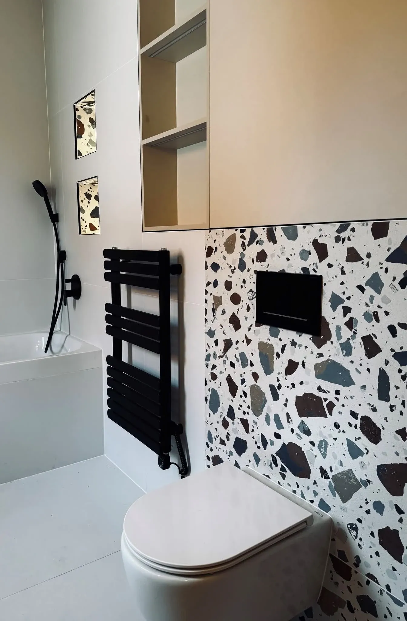 Fürdőszoba terrazzo mintás falburkolattal, fekete WC-nyomólappal és fekete törölközőszárítóval