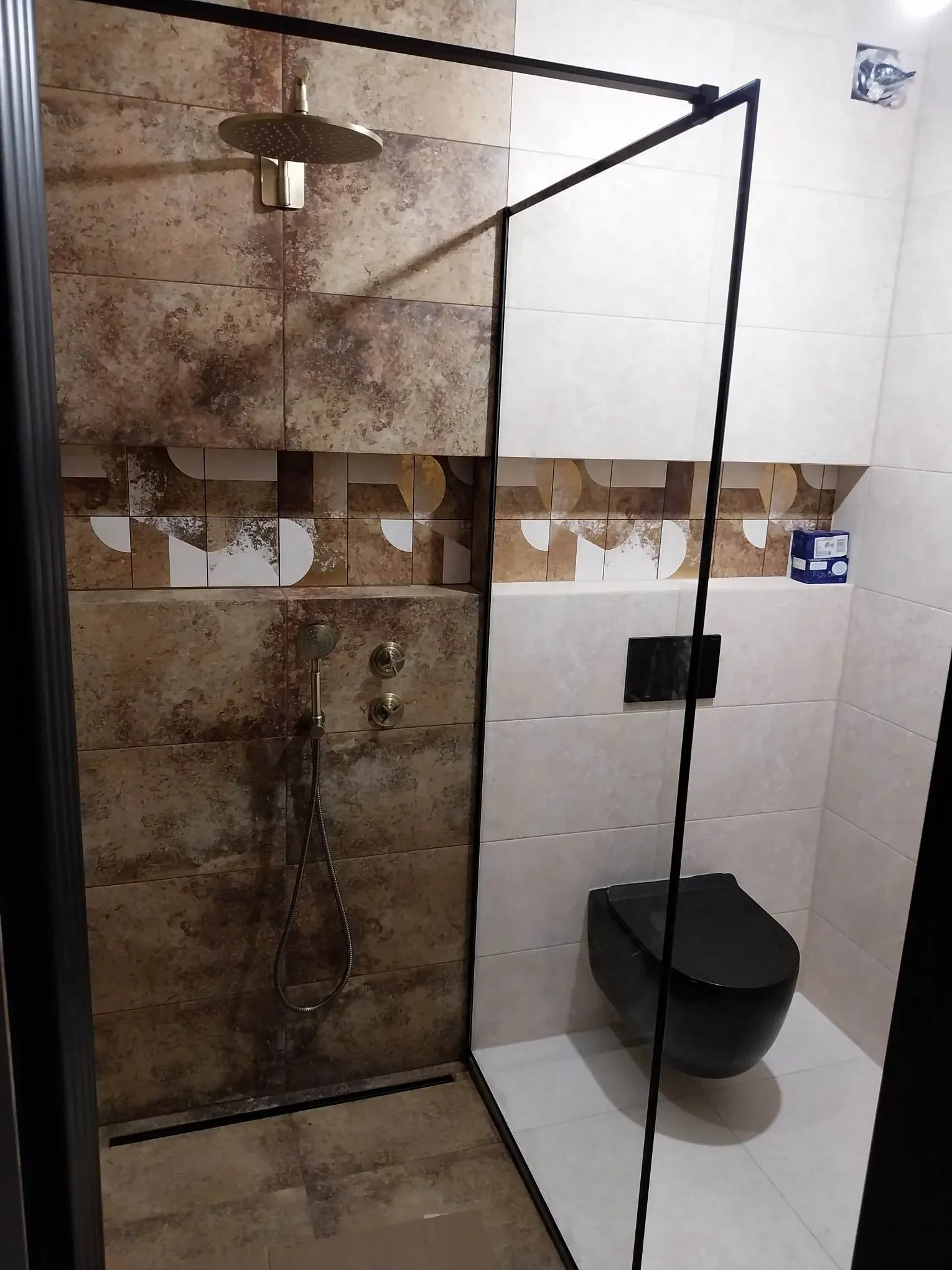 Zuhanyzó rozsda- és patinahatású burkolattal, arany színű zuhanyszettel és fekete fali WC-vel
